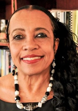 Belinda Biscoe has dark hair, pulled back, and is smiling. She is wearing a bold, black and white necklace and a black top. In the background are shelves with books.
